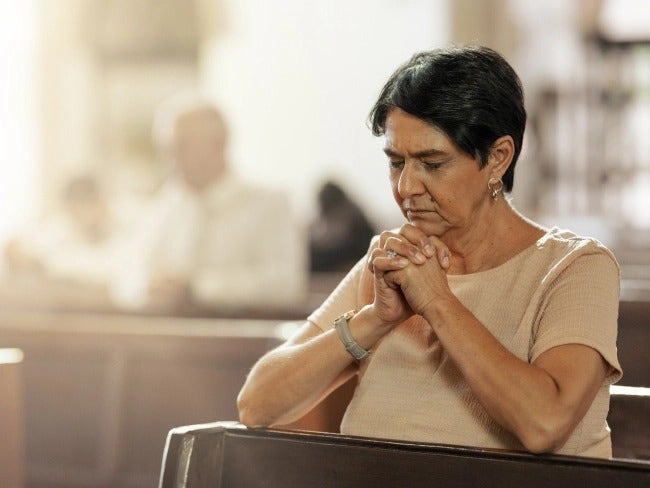 Una señora mayor con COVID-19 factores de riesgo está arrodillado en oración.