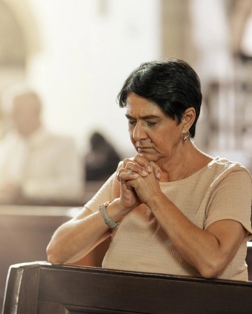 Una señora mayor con COVID-19 factores de riesgo está arrodillado en oración.